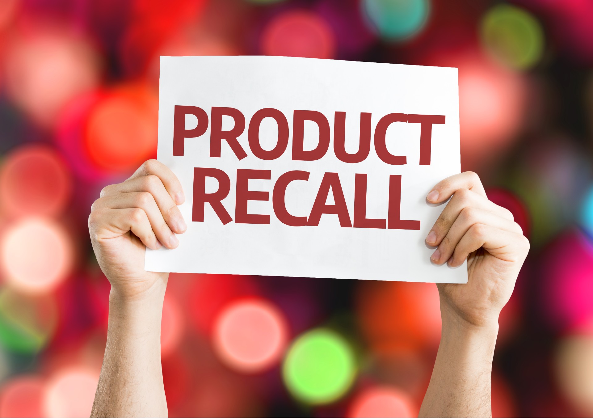 Closeup of a person's hands holding up a sign that reads "Product recall"; blurry colored lights in the background - baker ross toys